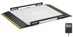 A flat, platform-style medical scale (seca XLine Base) with a smooth, white surface and yellow safety edges on the ramps. The scale has side railings and the brand name 'seca' is printed in the center. To the right of the platform, a small digital display unit (Basic Display) shows numerical data, connected by a cable.