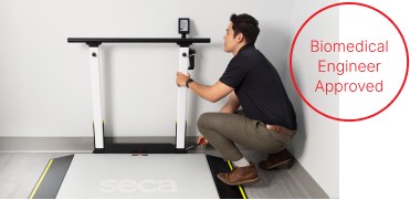 A biomedical engineer kneels next to a large digital weighing platform with safety edges and vertical support rails (seca XLine Hold). The engineer is performing maintenance or adjustments on the equipment, with a small toolkit visible nearby. A digital display is mounted at the top of the support rails. To the right of the image, a red circle contains the text 'Biomedical Engineer Approved.'