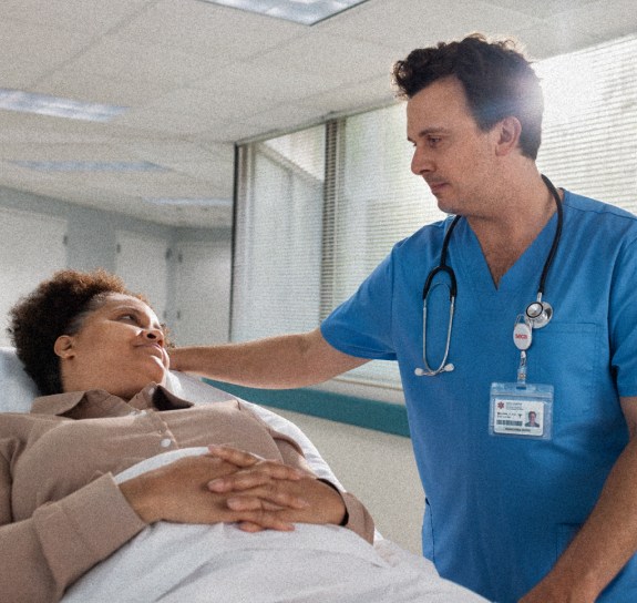 A healthcare professional in blue scrubs with a stethoscope gently places a hand on the shoulder of a patient lying in a hospital bed, offering comfort. The patient, a woman with short, curly hair, looks up at the healthcare worker, appearing calm and reassured. They are in a brightly lit hospital room with large windows in the background.