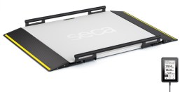 A flat, platform-style medical scale (seca XLine Base) with a smooth, white surface and yellow safety edges on the ramps. The scale has side railings and the brand name 'seca' is printed in the center. To the right of the platform, a small digital display unit (ID-Display™) shows numerical data, connected by a cable.