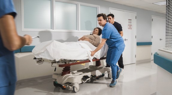 Two healthcare professionals, one in blue scrubs and the other in dark clothing, are moving a patient on a hospital stretcher down a brightly lit hallway. The patient, covered with a white blanket, lies calmly on the stretcher. A third medical professional is visible in the foreground, partially out of the frame, holding a clipboard. The hospital corridor is clean and modern, with windows and white walls.