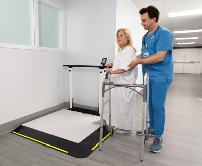 A nurse in blue scrubs assists a female patient standing on a large digital weighing platform with yellow safety edges (seca XLine Hold). The patient, wearing a hospital gown and holding a walker, stands on the scale with the nurse providing support from behind. The scene is set in a well-lit hospital hallway near a window, with medical equipment visible in the background.