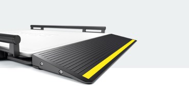 A close-up of the ramp section of a large medical weighing platform with a textured, black surface and a prominent yellow safety stripe along the edge (seca XLine Base). The ramp is slightly elevated and designed for smooth access onto the scale platform, which is partially visible in the background.