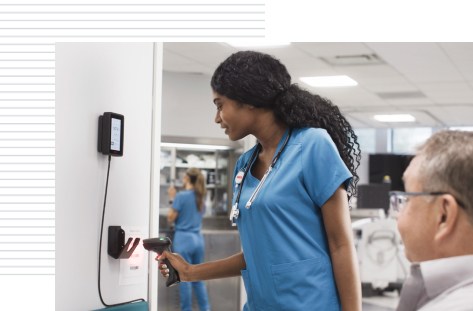 A nurse in blue scrubs scans a barcode using a handheld scanner on a wall-mounted device in a medical facility. The device displays information on a small digital screen. In the foreground, a patient in a wheelchair is partially visible, while in the background, medical equipment and other healthcare workers can be seen in the brightly lit room.