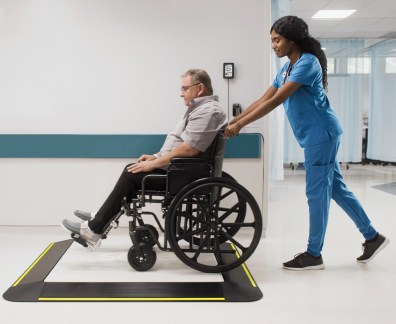 A nurse in blue scrubs is pushing a male patient in a wheelchair onto a large digital weighing platform with yellow safety edges (seca XLine Base), positioned in a hospital corridor. The patient is seated comfortably in the wheelchair, and the nurse is attentively guiding him onto the scale. The scene takes place in a clean, modern medical facility.